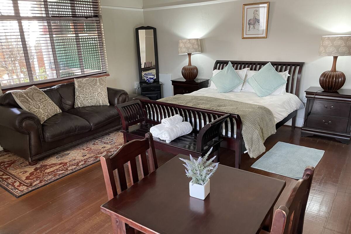 Un dormitorio bien amoblado con cama, mesitas de noche, cómoda, ventana con persianas, espejo y un rincón comedor con mesa y sillas.