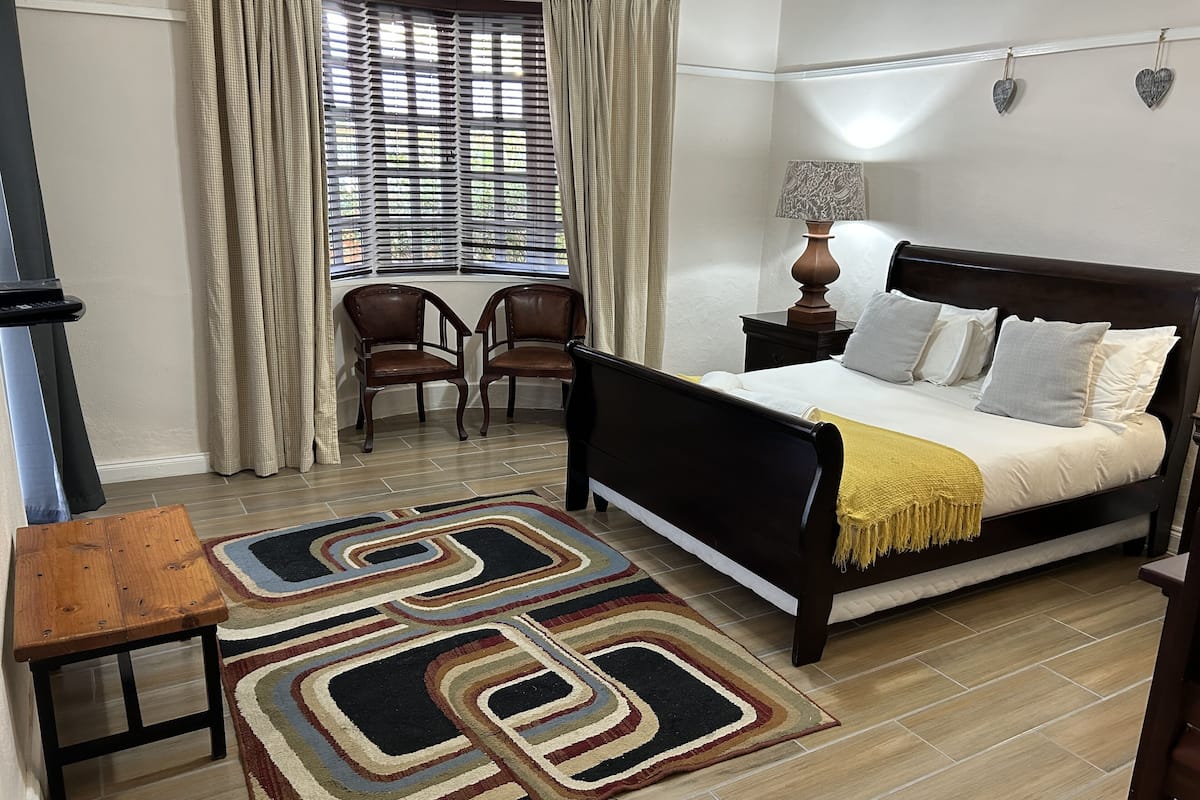Un dormitorio con cama, mesita de noche, lámpara, dos sillas, una ventana con cortinas y una alfombra estampada.