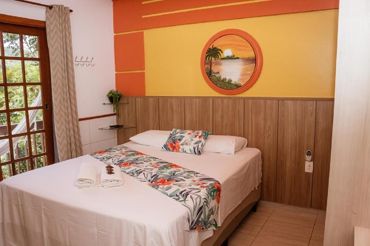 Ein ordentlich bezogenes Bett mit floralem Überwurf und zwei Kissen, ein Holzkopfende, ein an der Wand hängendes Bild von einem Sonnenuntergang über Wasser und ein Fenster mit Blick auf Grünflächen.