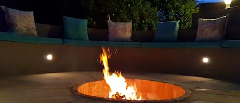 A circular fire pit with flames, surrounded by a low wall with cushions.