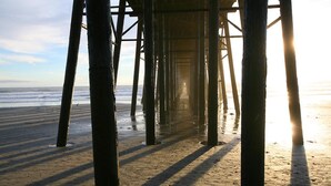 Una estructura de muelle con vigas de madera que se adentran en el agua, una playa de arena y el océano al fondo.
