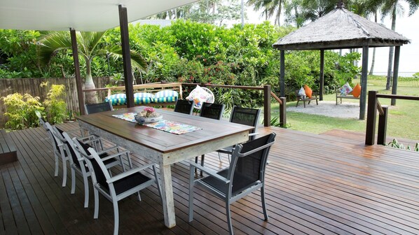 An outdoor dining area with a wooden deck, metal chairs, and a table set with a placemat and a bowl of flowers. - Absolute BeachFront Bliss @ Newell Beach (Newell)