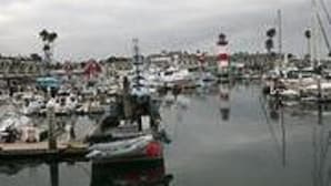 Un port de plaisance où sont amarrés de nombreux bateaux, un phare imposant et un ciel nuageux.
