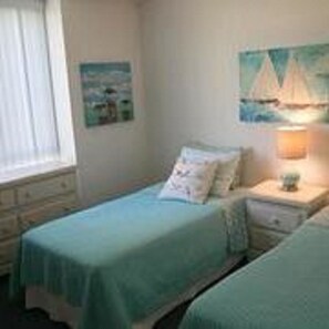 A bedroom with two single beds, a nightstand with a lamp, a dresser, and a painting of sailboats on the wall.