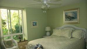 Quarto com cama, mesa de cabeceira, luminária, cadeira de vime, janela com vista para vegetação e ventilador de teto.