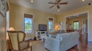 A spacious living room with a light blue sofa, wooden chairs, a television, and a ceiling fan.