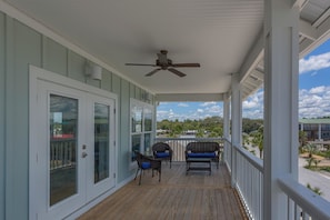 A covered porch with a ceiling fan, two wicker chairs with blue cushions, and a view of a residential area with houses and trees. - Mermaid Kisses (Port St. Joe)