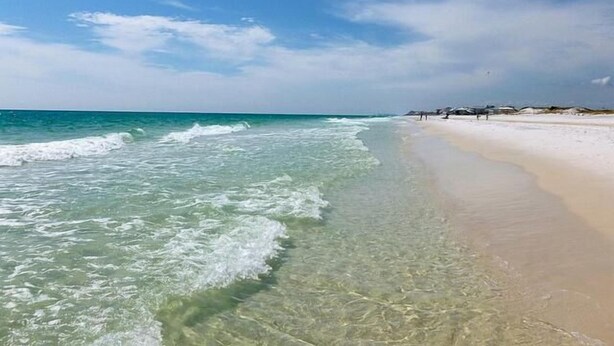 Una playa con agua cristalina, una orilla arenosa y algunas personas caminando.