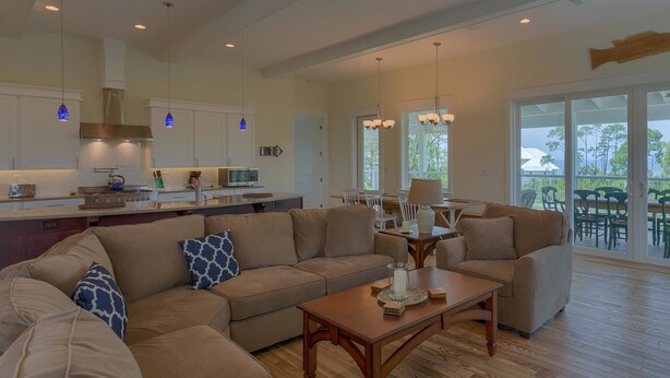 A spacious living area with a large beige sectional sofa, a wooden coffee table, and a dining table with chairs. A kitchen with white cabinets and a stainless steel range hood is visible in the background.