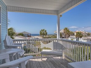 A wooden deck with white railings and chairs, overlooking a beach and residential buildings.