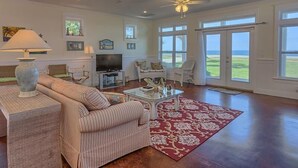 Un salon spacieux avec un canapé à rayures, une table basse et un tapis rouge à motifs. On y trouve un téléviseur posé sur un meuble, un ventilateur de plafond et de grandes fenêtres qui offrent une vue sur l’extérieur.