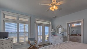 Une chambre à coucher comprenant un lit, une commode et une coiffeuse avec un miroir. Elle est dotée de grandes fenêtres et d’un ventilateur de plafond.