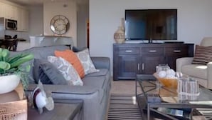 Una sala de estar moderna con un sofá gris, una mesa de centro y un televisor. Se ve un área de cocina al fondo.
