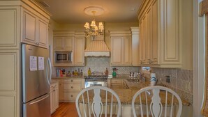 A kitchen with light-colored cabinets, a stainless steel refrigerator, a microwave, and a chandelier. There are two white chairs with patterned cushions at the kitchen island. - Maui Escape (Port St. Joe)