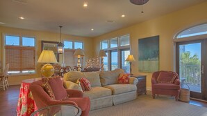 A spacious living room with a large sofa, armchairs, and a glass coffee table. There are multiple windows providing natural light and a view of the outdoors. - Maui Escape (Port St. Joe)