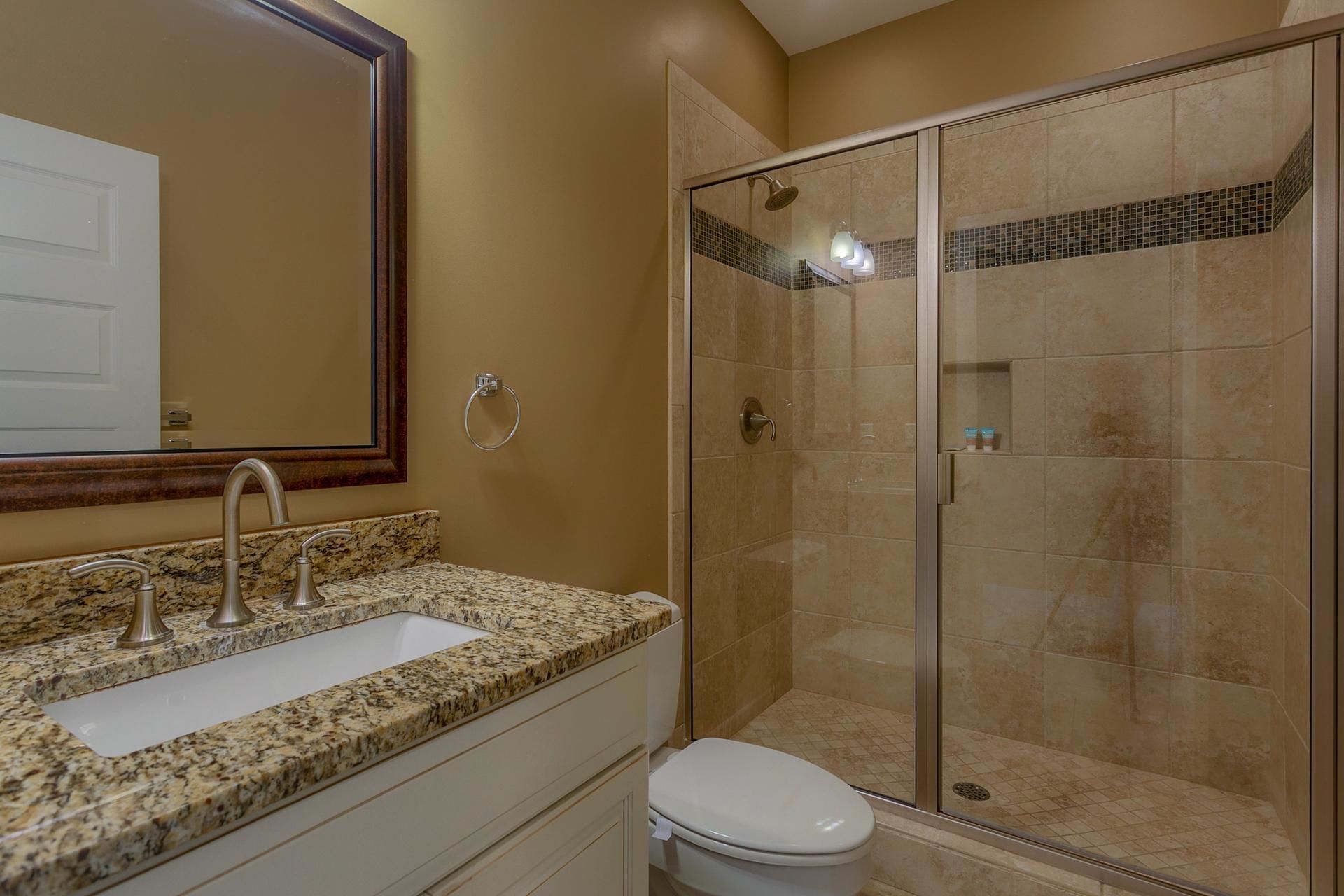 A bathroom with a granite countertop sink, a mirror, a toilet, and a glass-enclosed shower.
