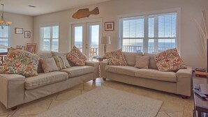 Ein Wohnzimmer mit zwei beigefarbenen Sofas, einem Deckenventilator und Blick auf den Ozean durch große Fenster.