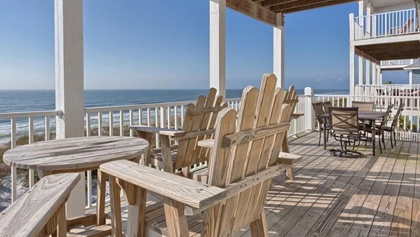 Eine Holzterrasse mit Adirondack-Stühlen und einem Tisch mit Blick auf den Ozean.
