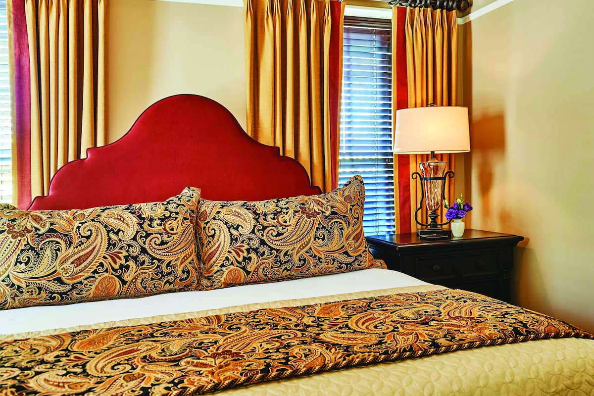 A hotel room with a red headboard, patterned bed linens, a bedside table with a lamp, and curtains.