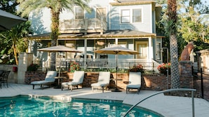 A pool area with lounge chairs and umbrellas, a brick wall, and a two-story building with a balcony in the background.