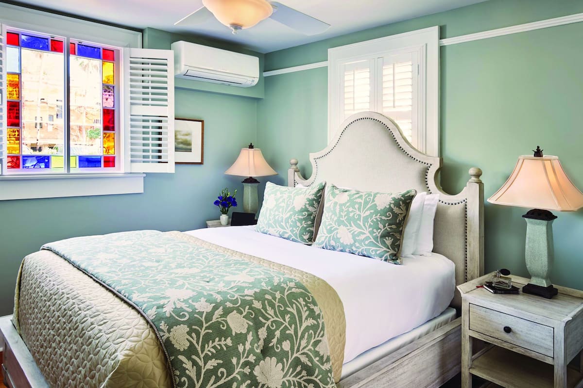 A bedroom with a bed, two bedside tables, a window with shutters, and an air conditioning unit.