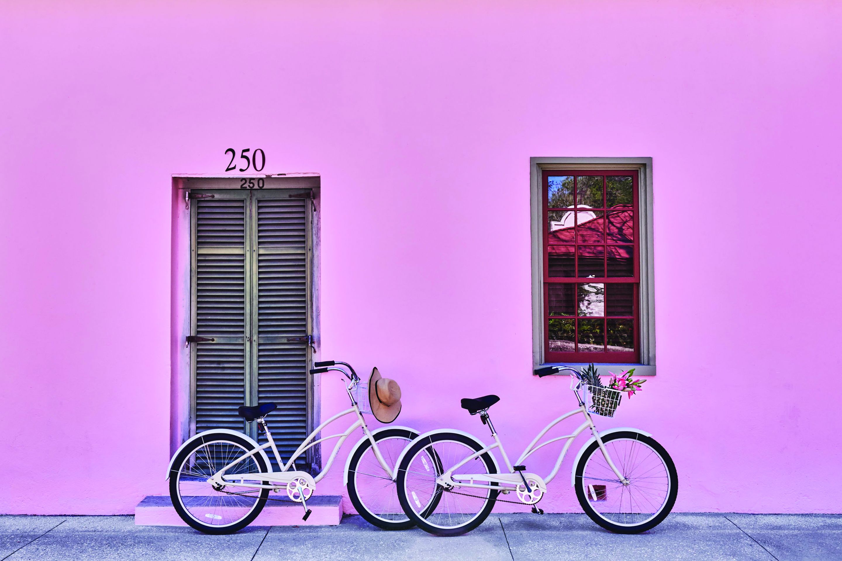 Uma bicicleta branca com um chapéu e uma cesta em frente a uma parede rosa, ao lado de uma porta com a inscrição '250'.