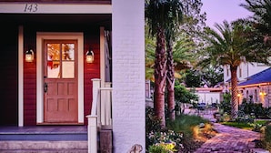 A wooden house with a red door and white trim, a stone statue, and a paved pathway leading to a patio area.
