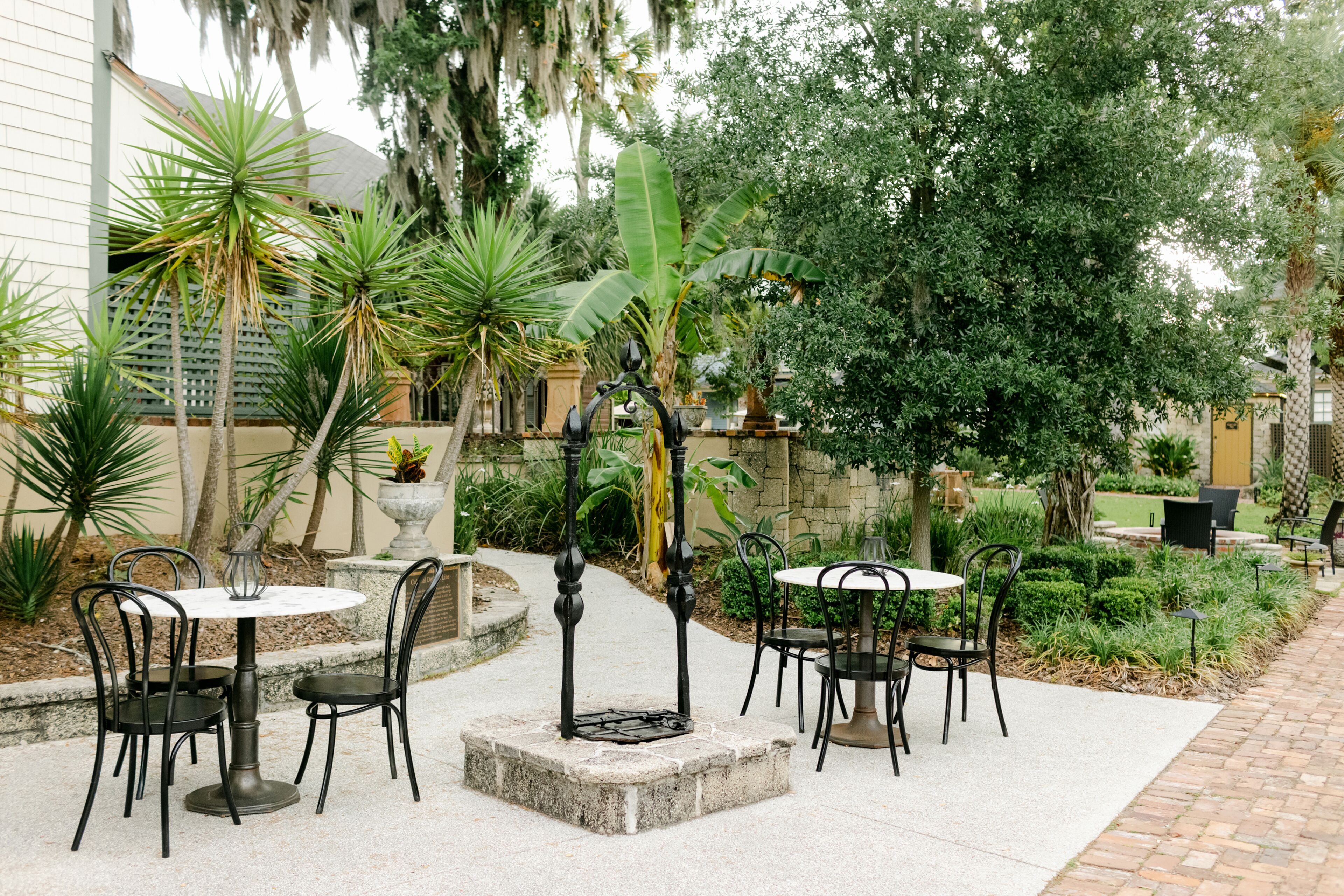 Un patio al aire libre con mesas y sillas redondas, rodeado de abundante follaje y un muro de piedra.