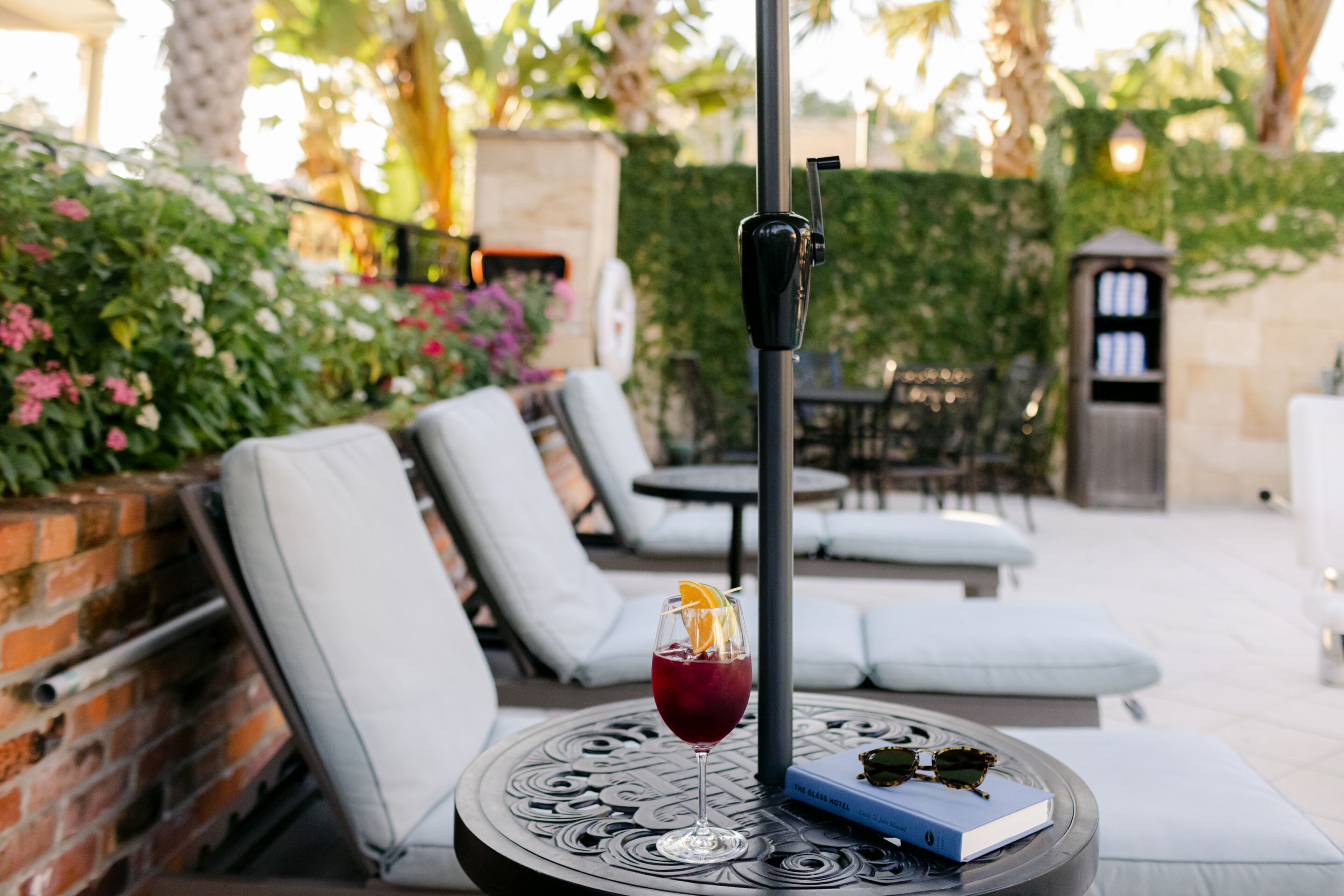Un vaso de bebida roja con una rodaja de naranja sobre una mesa redonda, un libro y un poste negro en un patio con sillones de descanso y un muro de ladrillo.