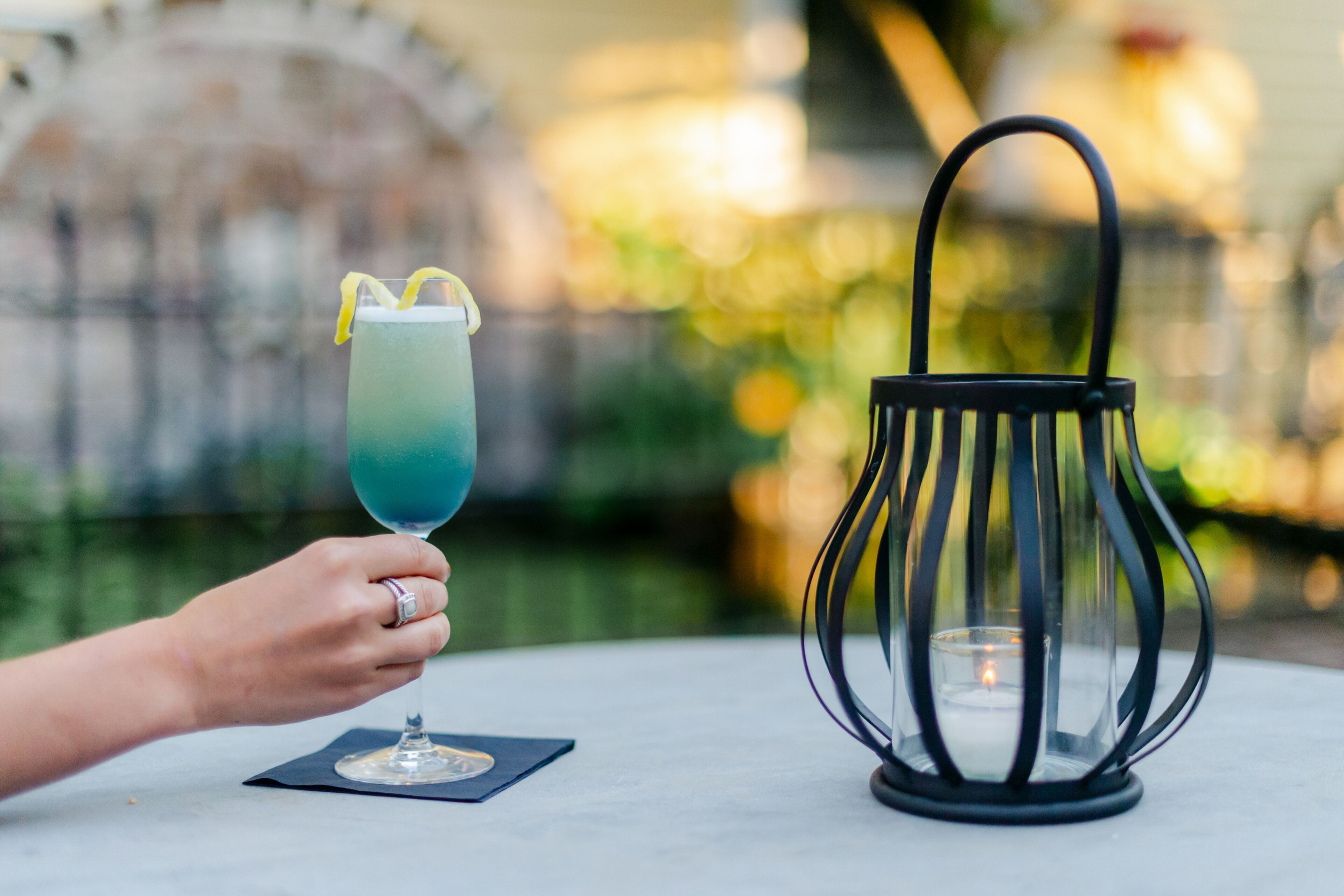 A hand holding a glass with a blue drink and a lemon slice, next to a black lantern on a table.
