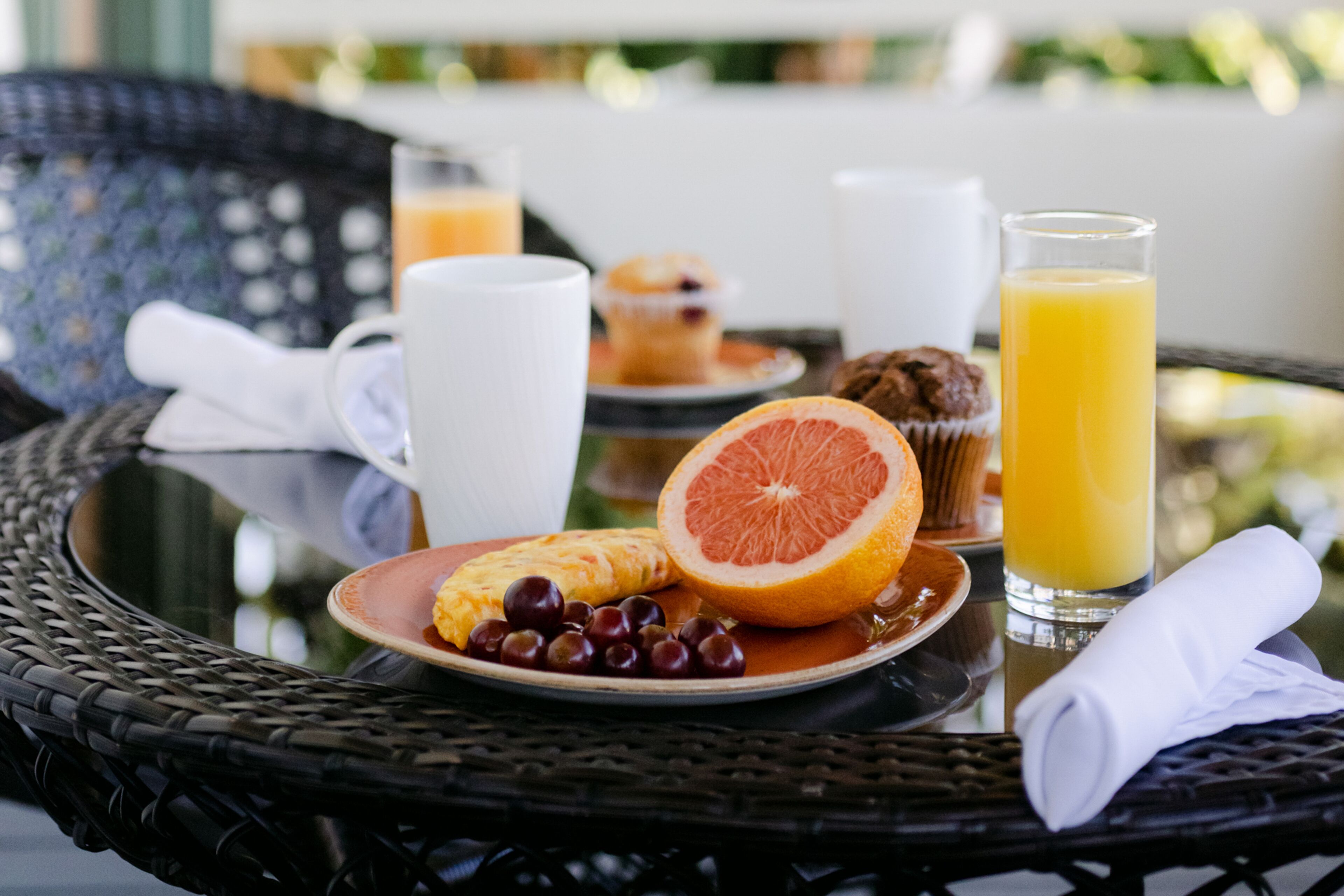 Une table en osier dressée pour le petit-déjeuner, avec une assiette de nourriture, un verre de jus d’orange et un muffin.