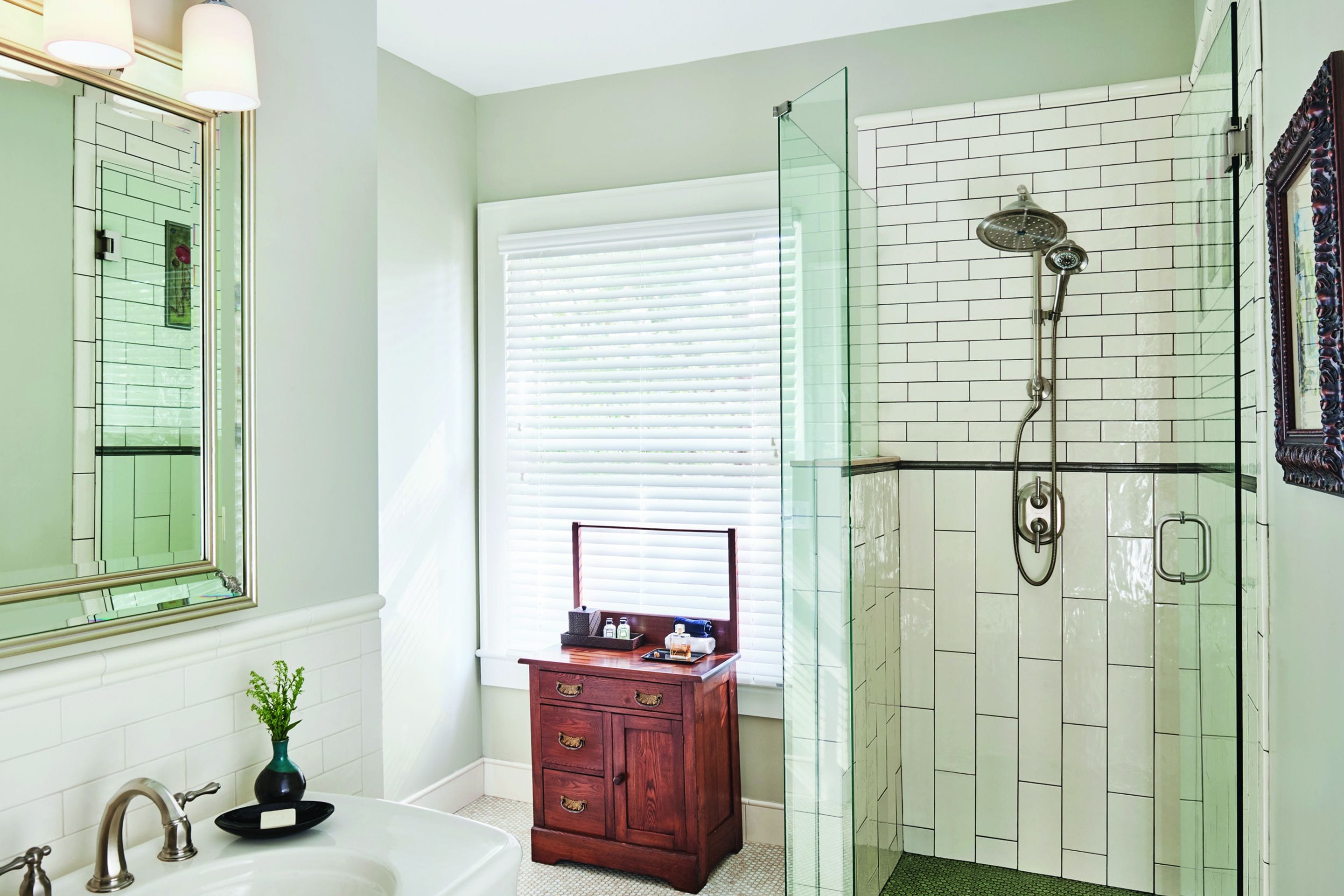 Une salle de bain équipée d’un meuble-lavabo, d’un miroir, d’une douche avec une porte en verre et d’une fenêtre avec des stores.