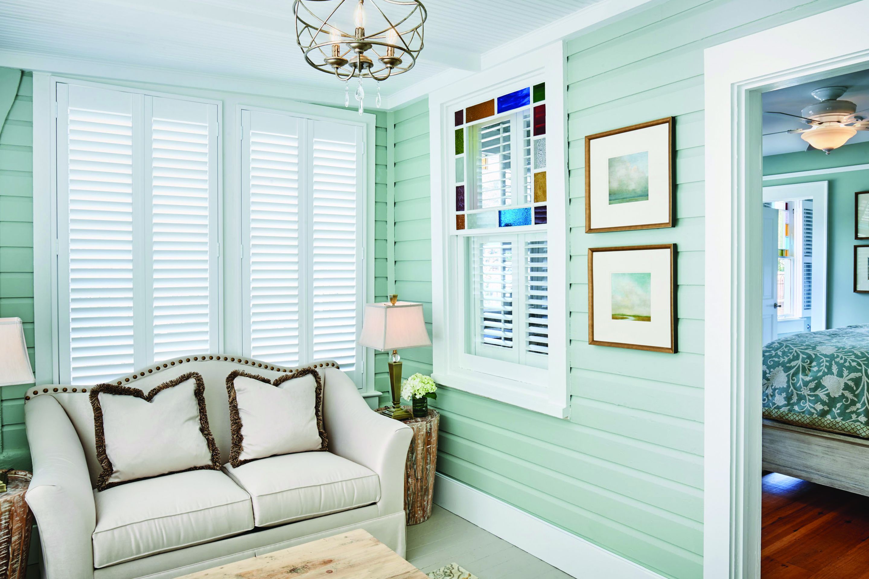 A room with a light green wall, white shutters, a white sofa with brown trim, a wooden coffee table, and a chandelier.