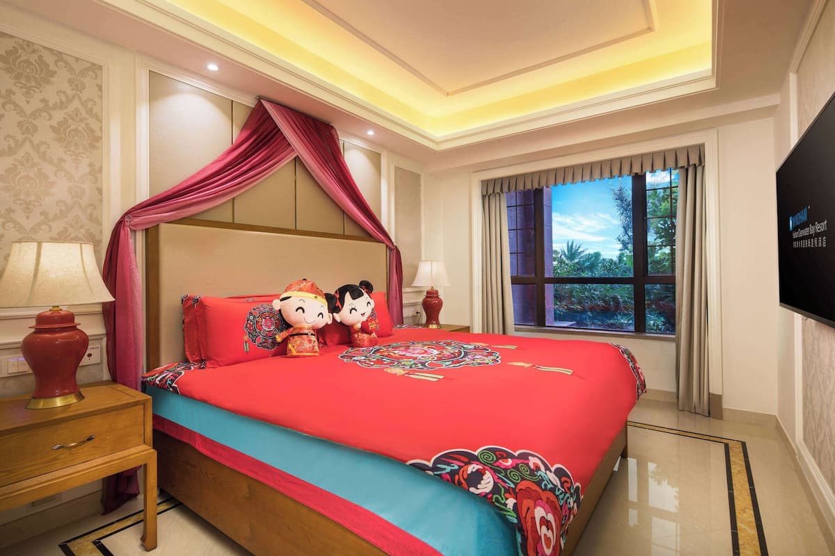 Habitación de hotel con una cama grande, una mesita de noche, una lámpara, una ventana con cortinas y un televisor montado en la pared.