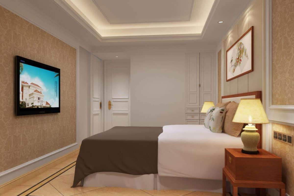 Una habitación de hotel con una cama, una mesita de noche con una lámpara, un televisor en la pared y una puerta que da a otra habitación.