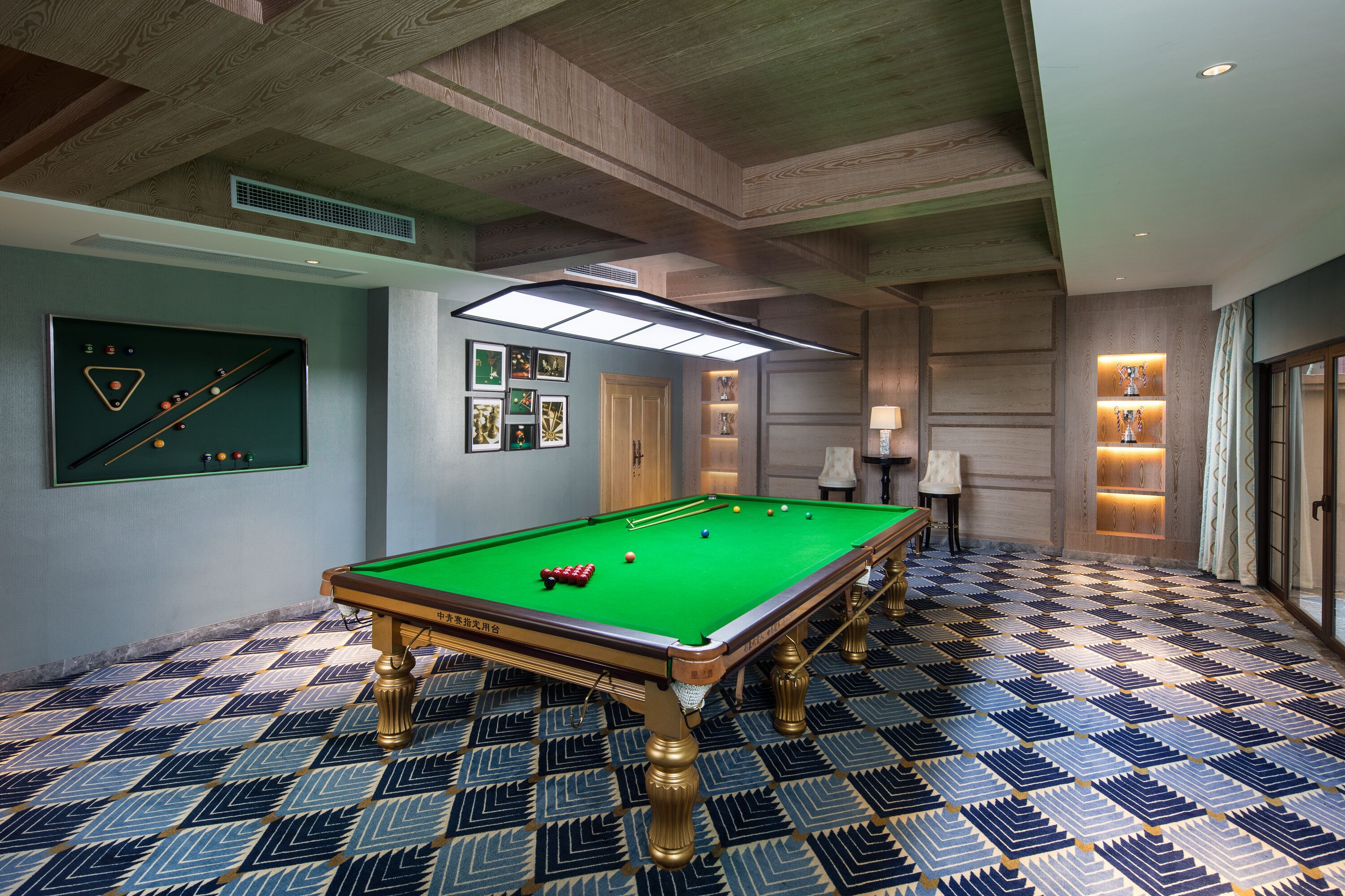 A pool table in a room with patterned flooring, a ceiling with wooden beams, and a wall-mounted billiards table.