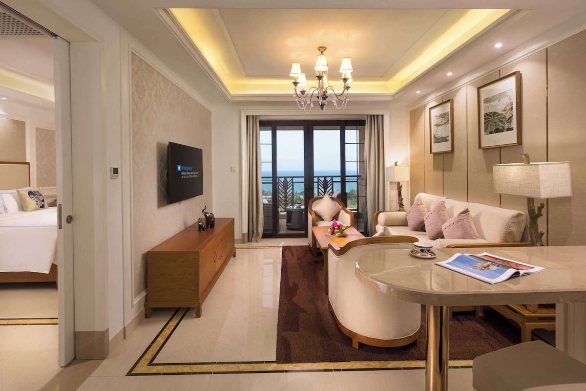 Una habitación de hotel moderna con una cama grande, una zona de estar con sofá y sillones, una mesa de comedor, un televisor y un balcón con vistas.