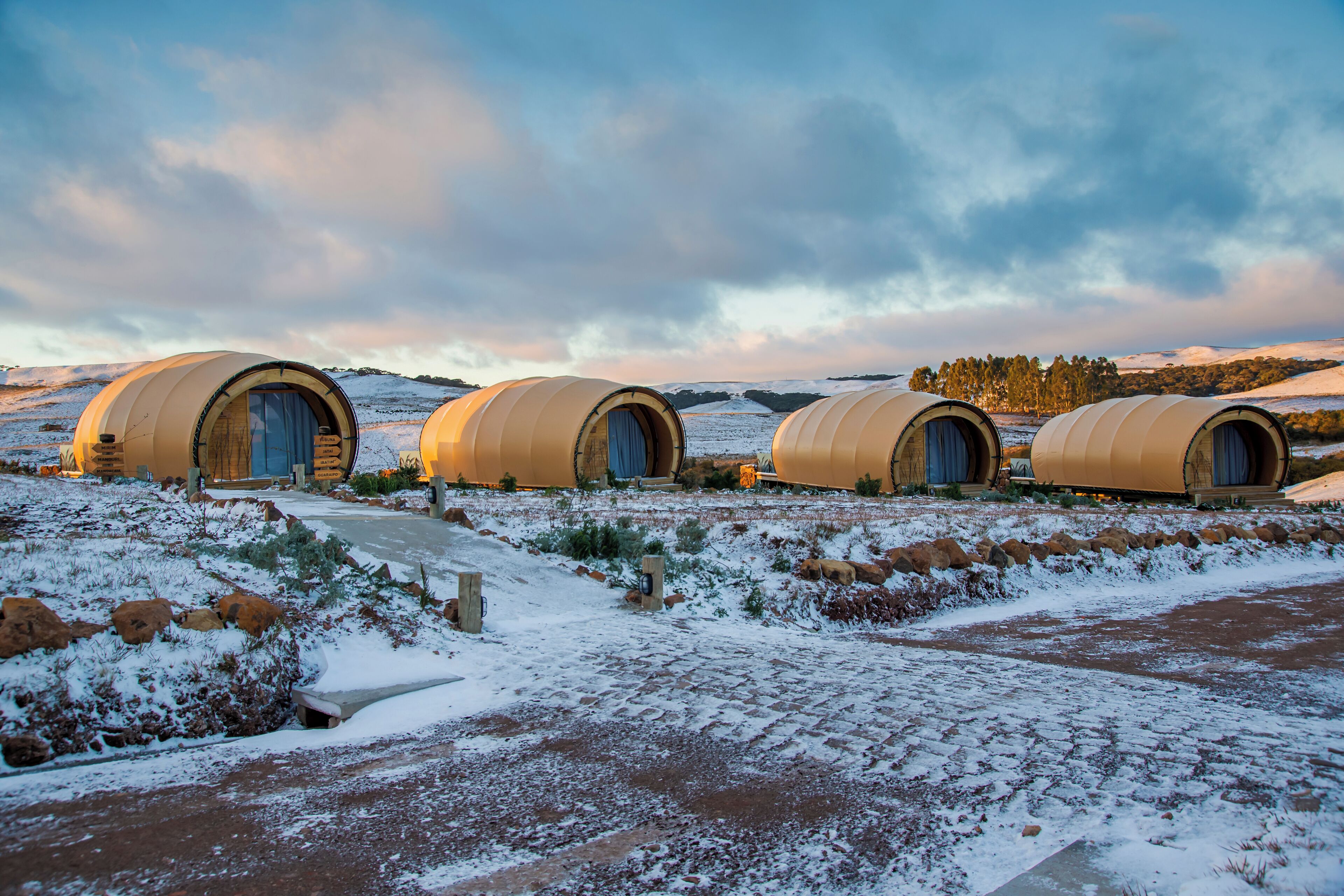 Cuatro estructuras redondas con techos color beige y puertas azules, ubicadas en un paisaje nevado con cielo despejado.