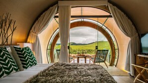 A bedroom with a large circular window offering a view of a green landscape. There is a bed with a furry rug, a television on a wooden stand, and a table with two glasses of wine. - Parador Cambará do Sul (Cambara do Sul)