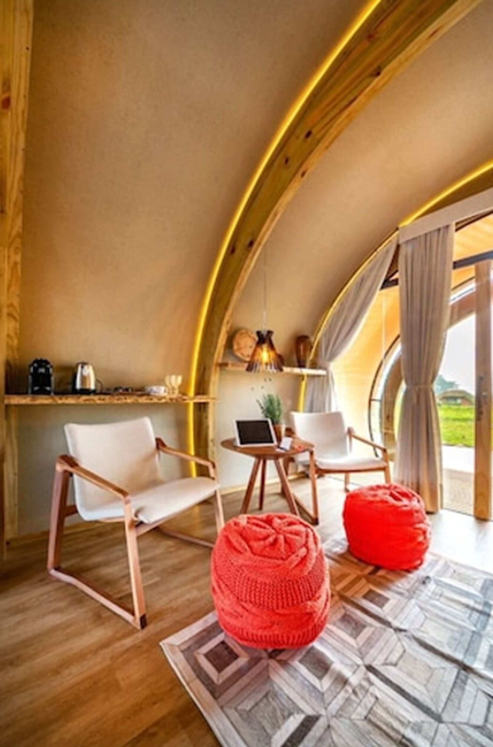 A cozy interior with a curved ceiling, wooden flooring, a red knitted pouf, and a laptop on a small table.