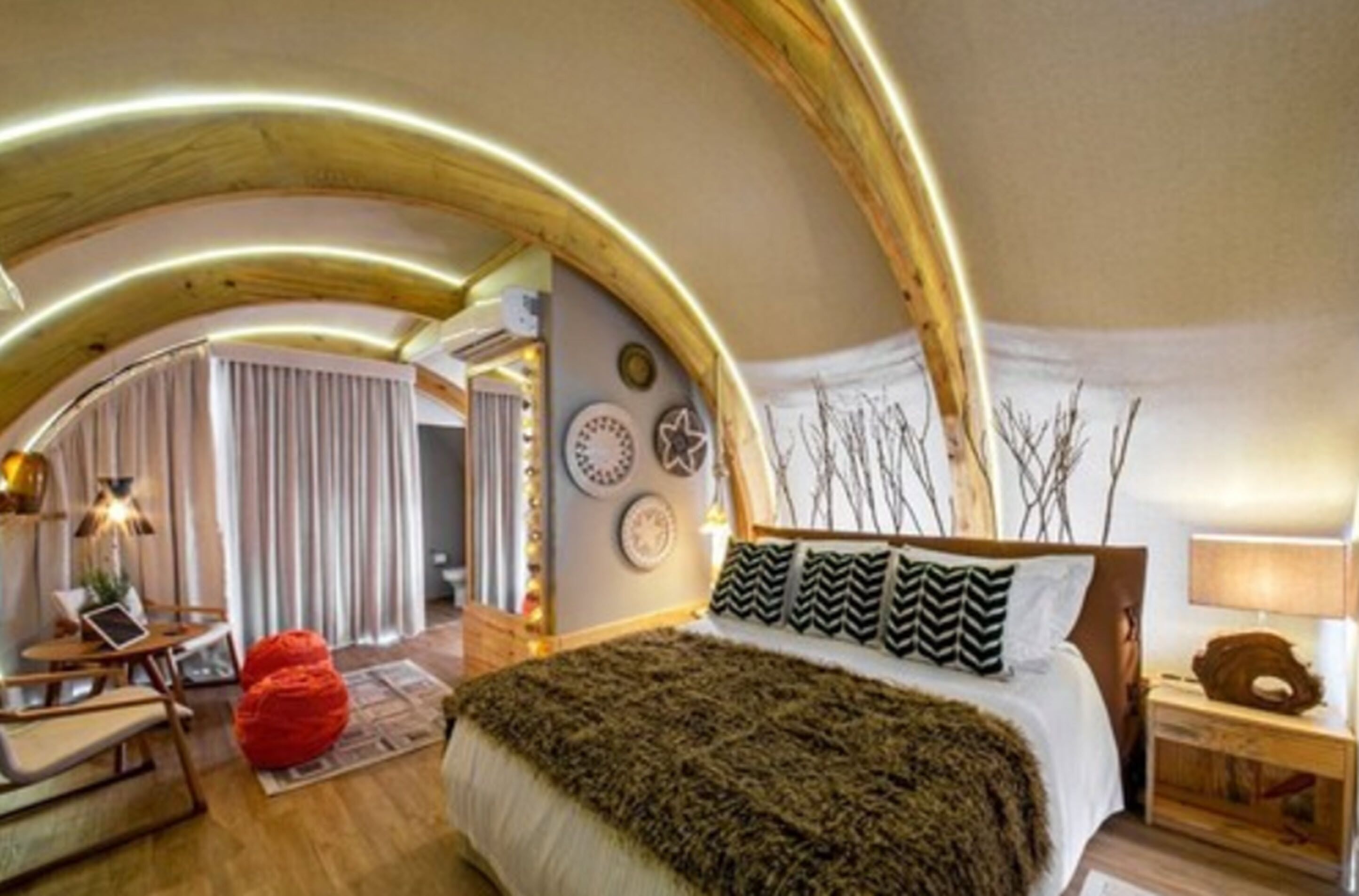 A cozy bedroom with a curved ceiling, a bed with patterned pillows, a red bean bag, and a bedside table with a lamp.