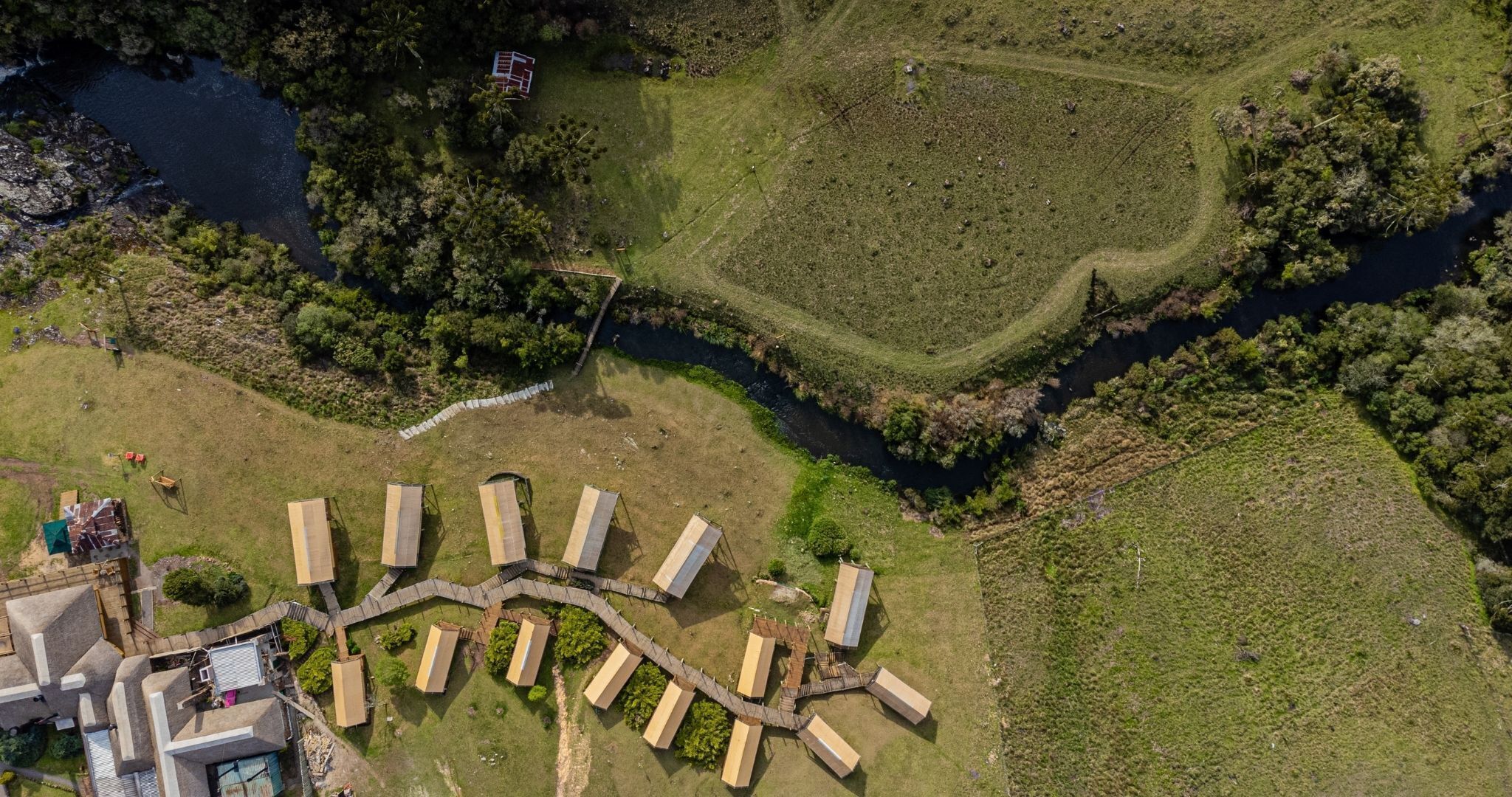 An aerial view of a campsite with tents arranged in a semi-circle, a small building, and a river.