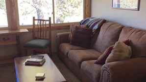 A living room with a brown sofa, a wooden coffee table, a green chair, and a window with a view of trees. - Star Mesa 2 Bedroom Holiday Home By Pinon Vacation Rentals (Howard)