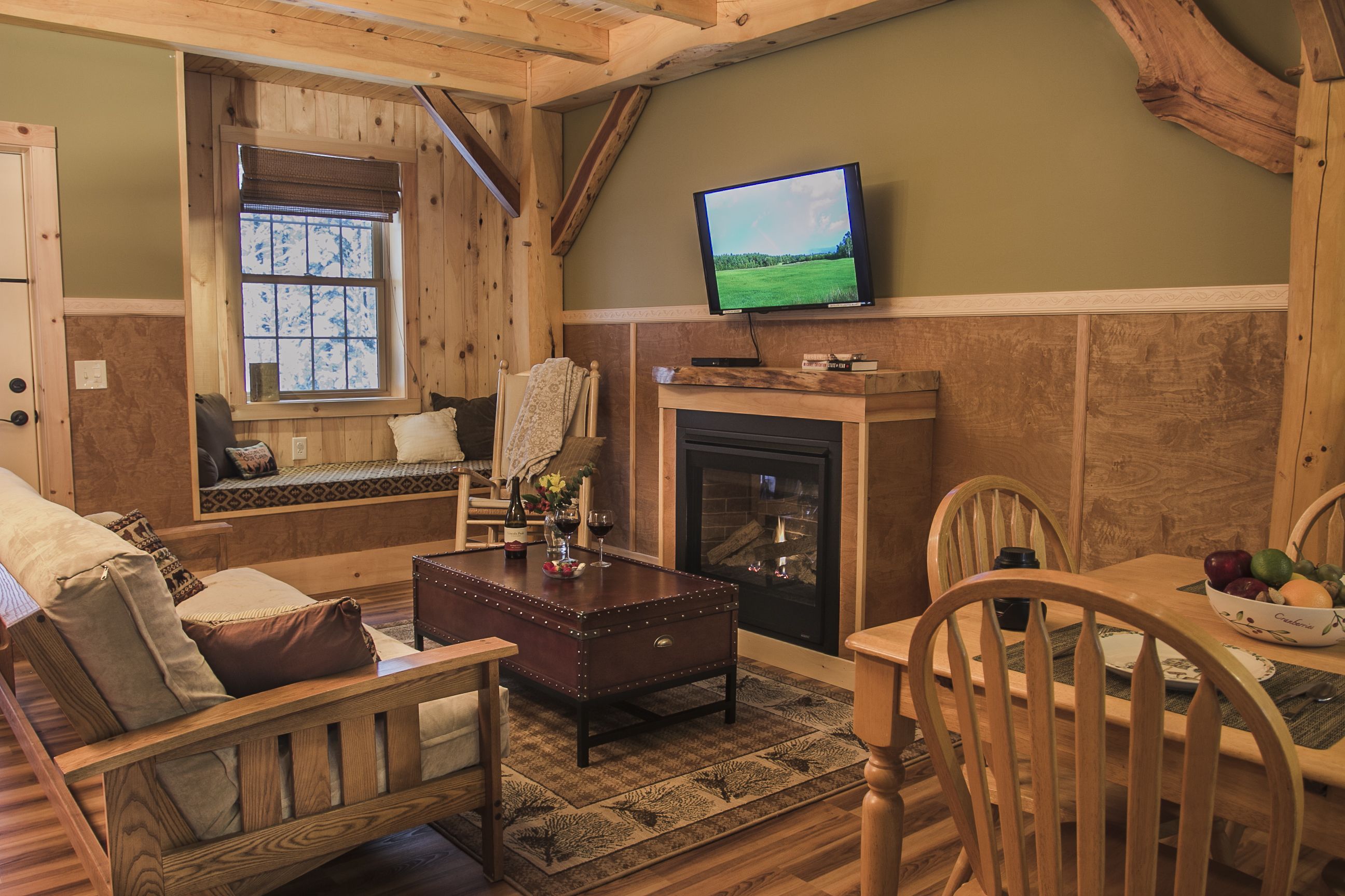 A cozy living room with a wooden bench, a fireplace, a television, and a dining area with a bowl of fruit.