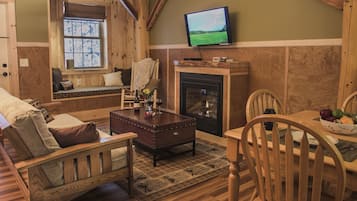 A cozy living room with a wooden bench, a fireplace, a television, and a dining area with a bowl of fruit.