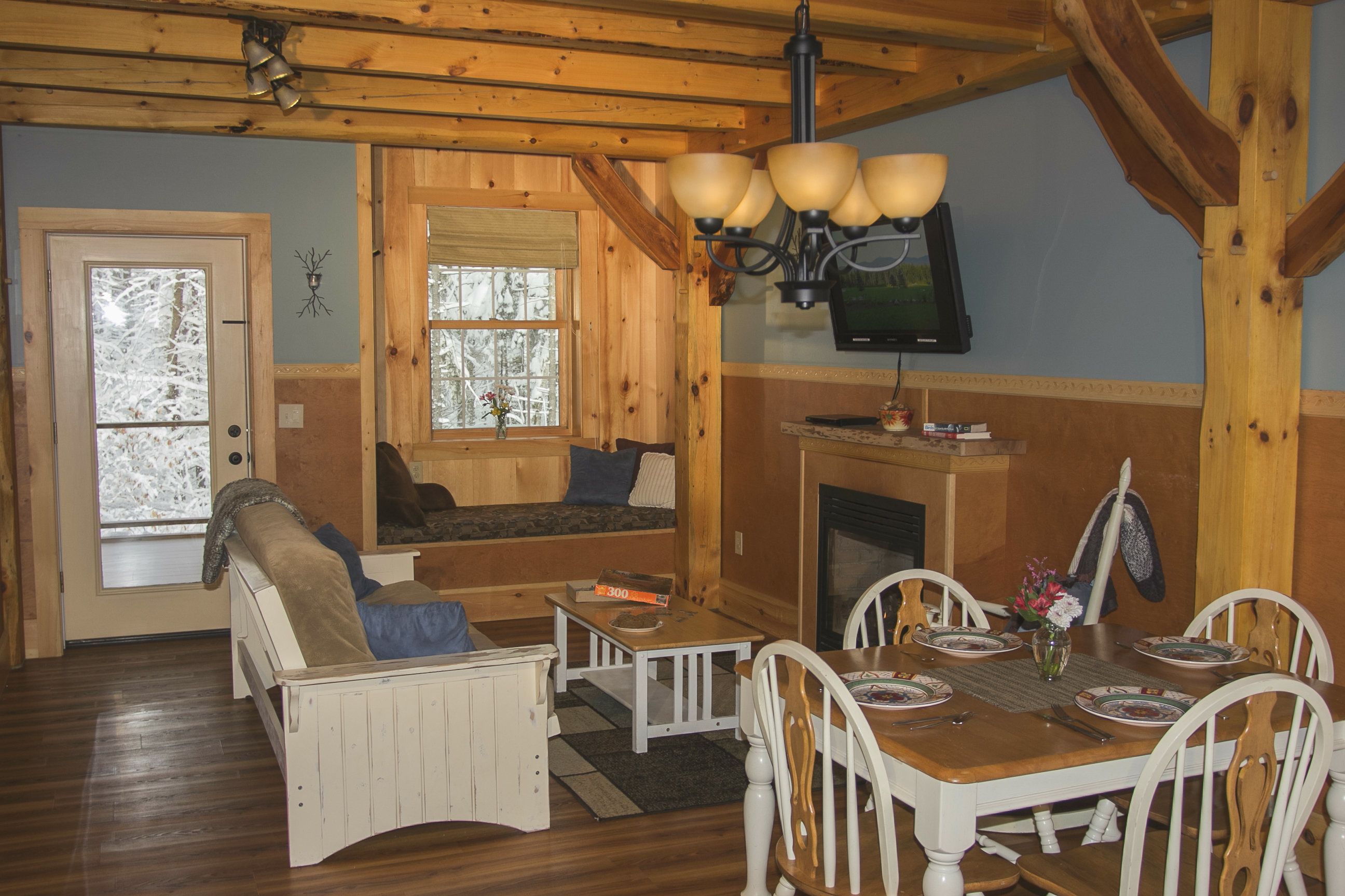 A cozy cabin interior with a wooden dining table, white chairs, a sofa, and a fireplace.