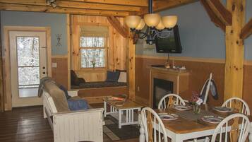 A cozy cabin interior with a wooden dining table, white chairs, a sofa, and a fireplace.
