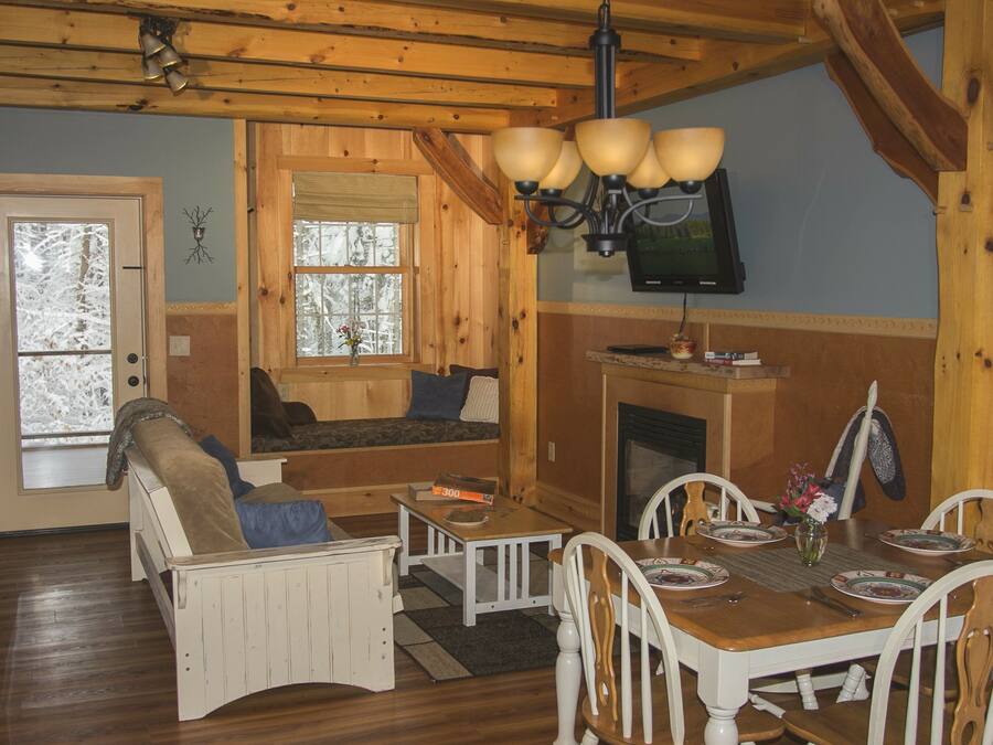 A cozy cabin interior with a wooden dining table, white chairs, a sofa, and a fireplace.