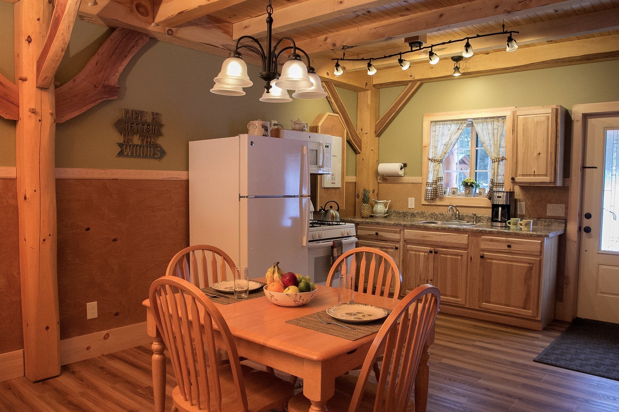 Una mesa de comedor de madera con cuatro sillas, un refrigerador blanco y un microondas en una cocina con armarios de madera y un ventanal con cortinas.