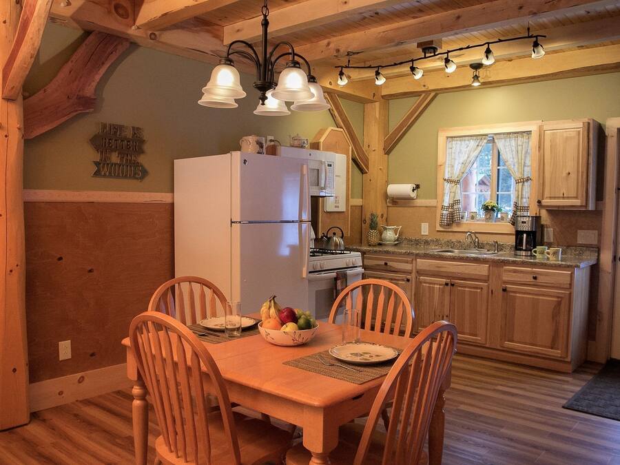 Una mesa de comedor de madera con cuatro sillas, un refrigerador blanco y un microondas en una cocina con armarios de madera y un ventanal con cortinas.